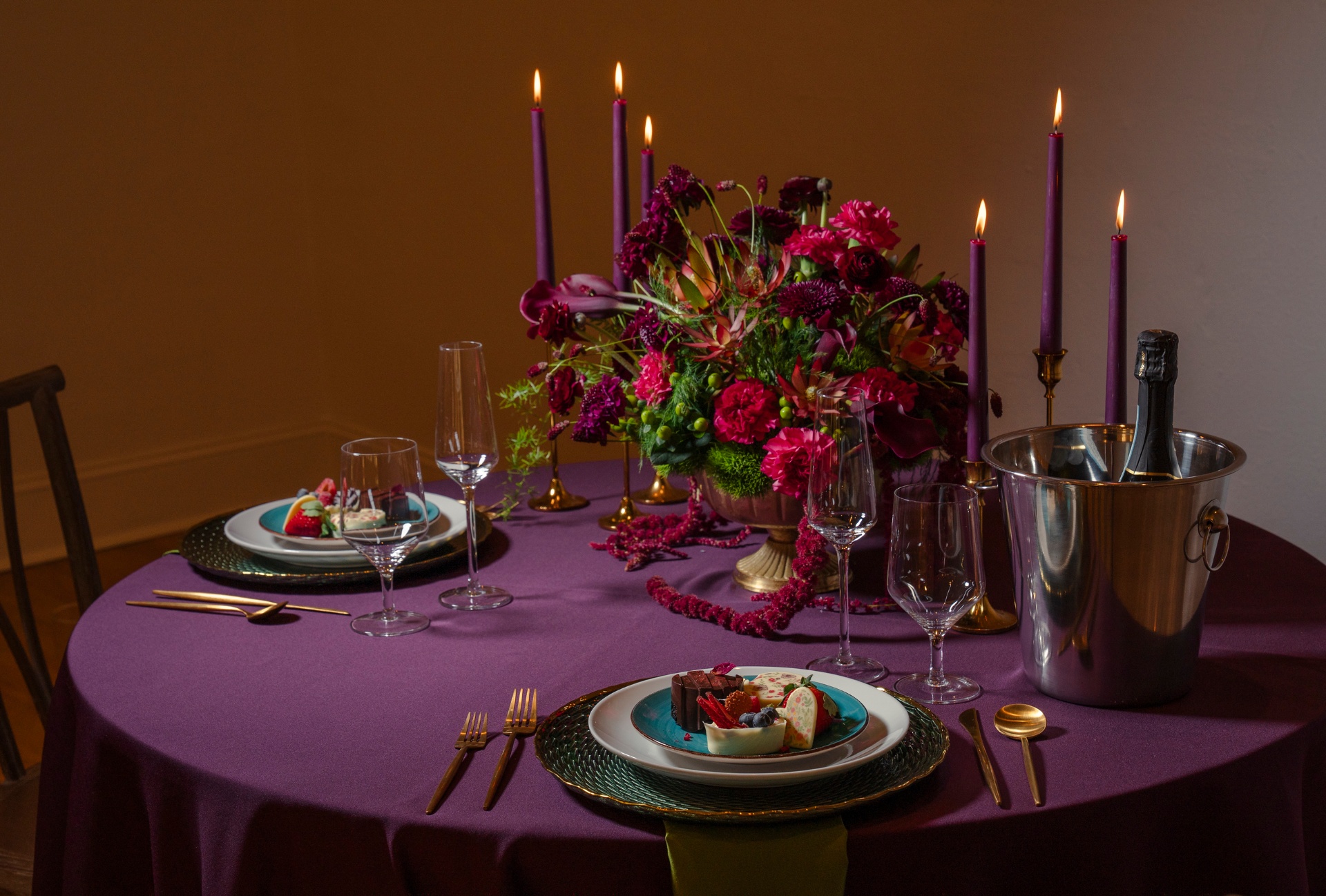 Plum linen round table set for two with crimson floral urn, taper candles and champagne on ice