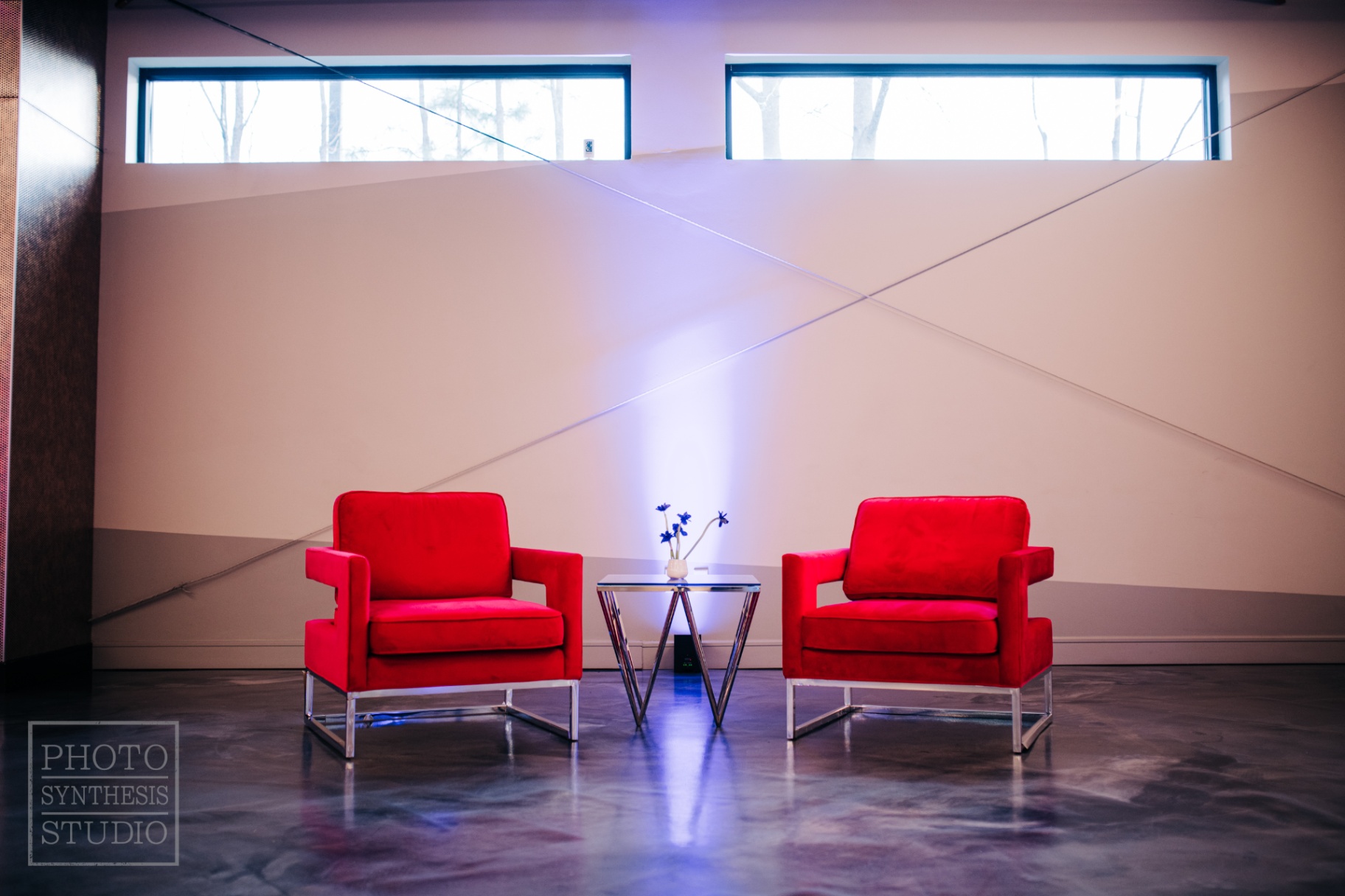 Symmetrical pair of red velvet chairs framing a chrome side table on polished concrete