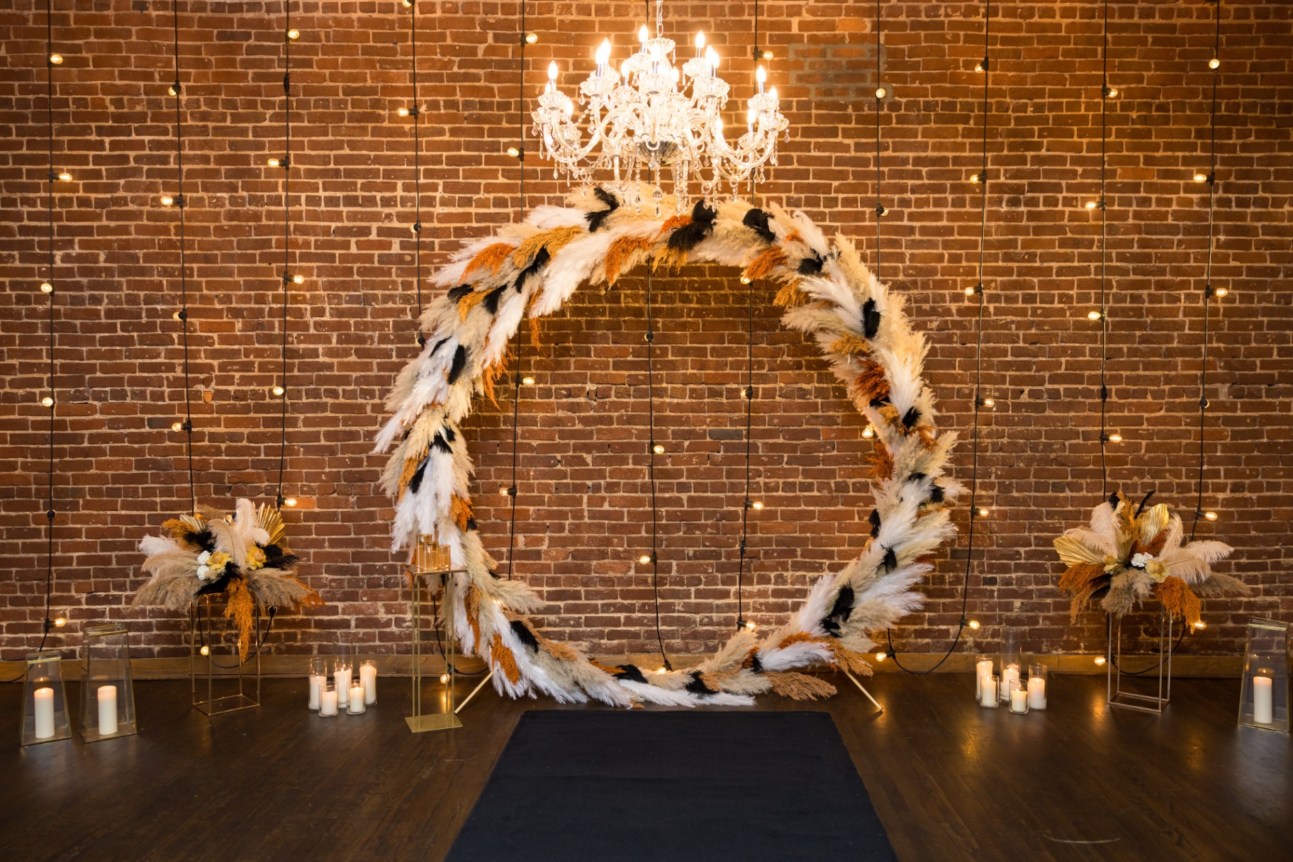 Oversized circular feather and pampas halo backdrop under a crystal chandelier on exposed brick