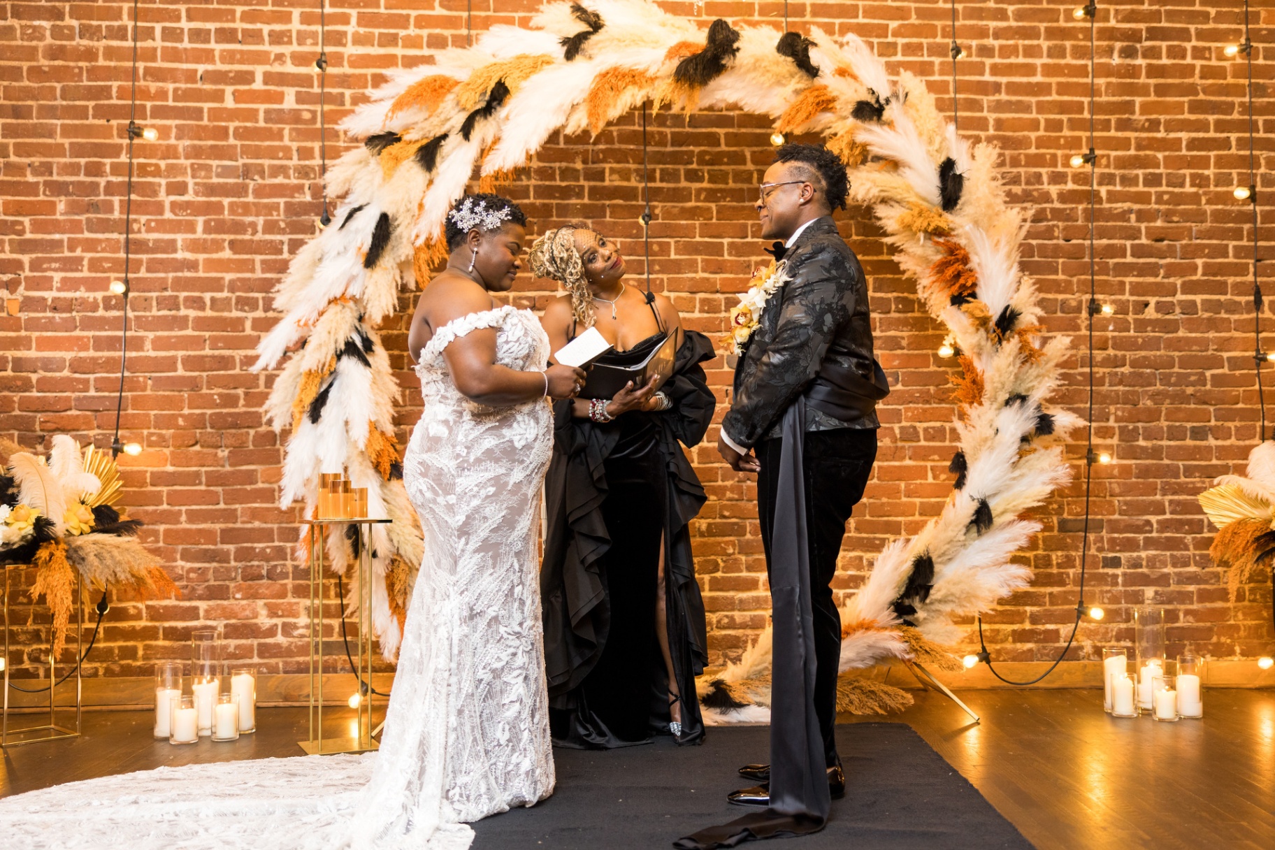 Brides exchanging vows beneath a feathered ring arch in a candlelit brick loft